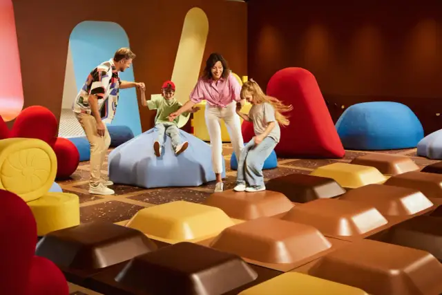 Rodzina bawi się w Muzeum Czekolady Warszawa, skacząc po gigantycznych kostkach czekolady i kolorowych pufach.
