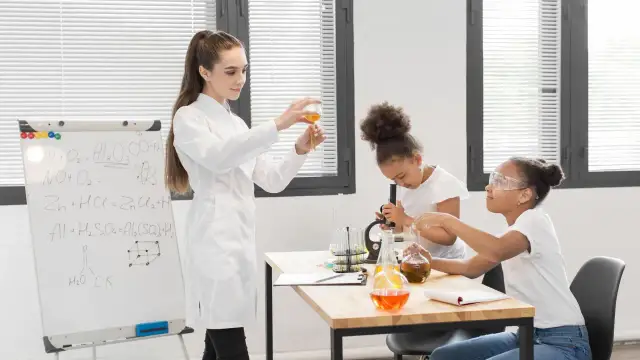 Nauczycielka demonstruje doświadczenie chemiczne, podczas gdy uczniowie pracują z mikroskopem i probówkami. Na tablicy widać wzory chemiczne, pomocne przy konkursy przedmiotowe arkusze.