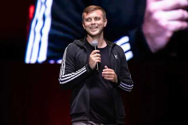 Uśmiechnięty Krajewski na scenie, trzyma mikrofon. Jego stand up rozgrzewa publiczność.