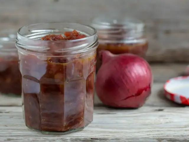 Słoik z konfiturą z czerwonej cebuli, obok leży czerwona cebula i pokrywka.