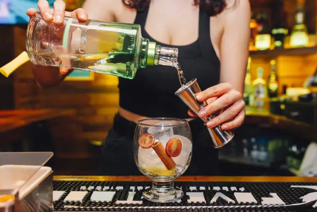 Barman nalewa alkohol z butelki do miarki nad kieliszkiem z lodem i owocami.