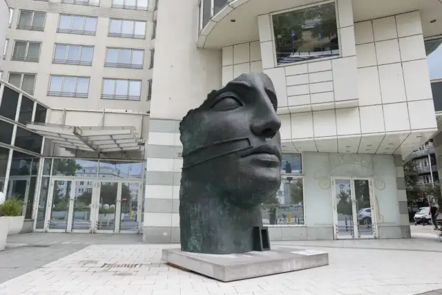 Okaleczona, ciemna rzeźba głowy z opaską na ustach, na tle nowoczesnego budynku.