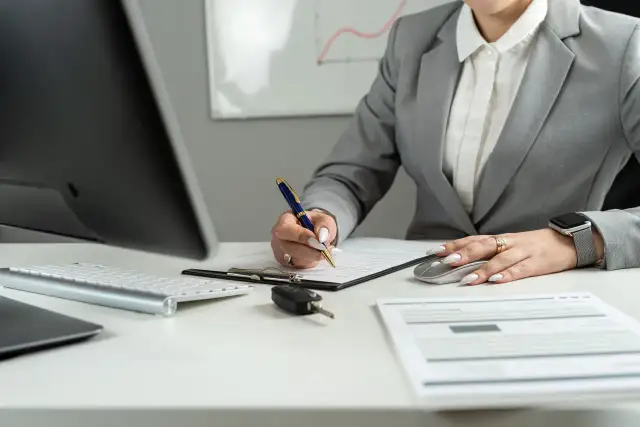 Kobieta w garniturze podpisuje dokumenty, obok kluczyki do auta i myszka. Zastanawia się, ile za km delegacji.