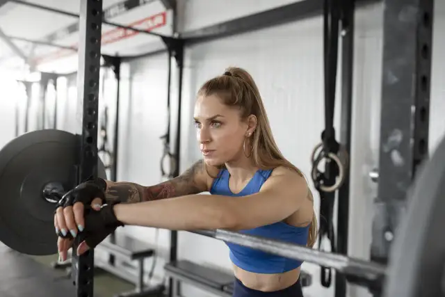 Kobieta w rękawiczkach opiera się o sztangę. To może być część jej treningu FBW. Co to jest?