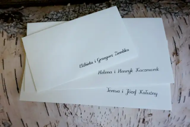 Białe koperty ślubne z elegancko wypisanymi nazwiskami gości na tle kory drzewa.