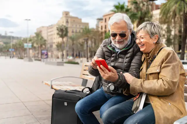 Uśmiechnięta para seniorów na ławce w mieście przegląda telefon. Czyżby sprawdzali, co to roaming, planując kolejną podróż?