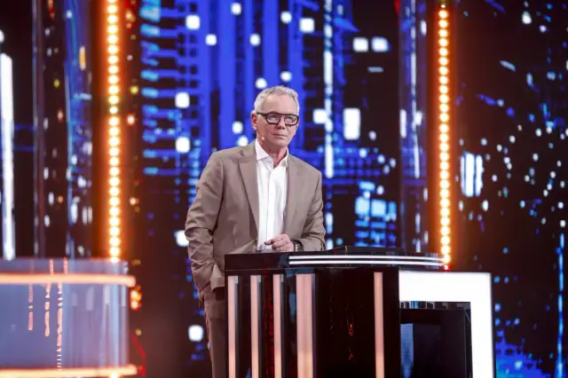 Mężczyzna w garniturze stoi przy pulpicie na tle sceny z niebieskimi światłami.