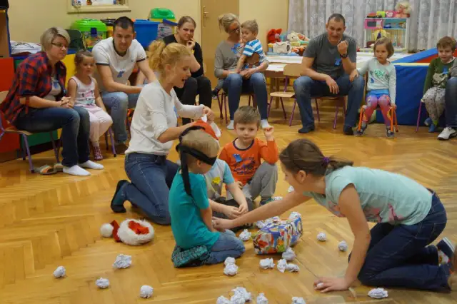 Zajęcia integracyjne z rodzicami w przedszkolu: dzieci z zawiązanymi oczami szukają prezentu, rodzice obserwują.