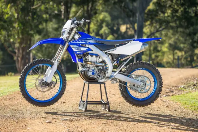 Niebiesko-biały motocykl Yamaha WR450F na stojaku, na tle drzew i ziemi.