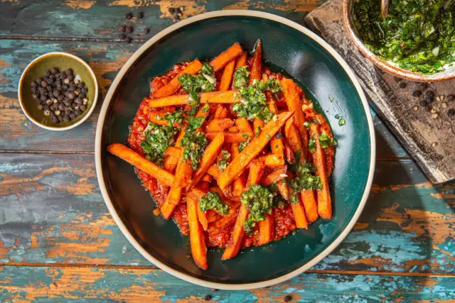 Pomarańczowe frytki z batatów podane z sosem pomidorowym i posypane zieloną, ziołową posypką. Obok miseczka z pieprzem.