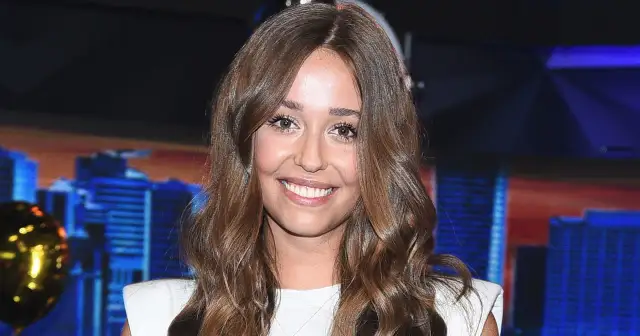 Uśmiechnięta prezenterka, która prowadzi koło fortuny z Brzozowskim, w białej sukience na tle miasta.