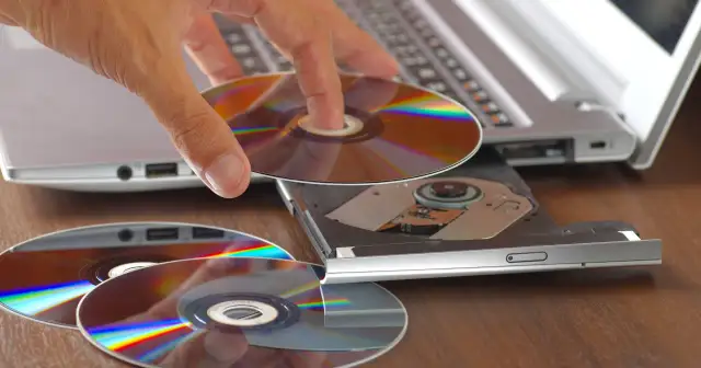 Dłoń wkłada płytę do napędu laptopa, przygotowując się do nagrania danych. Program do wypalania płyt jest kluczowy do archiwizacji.