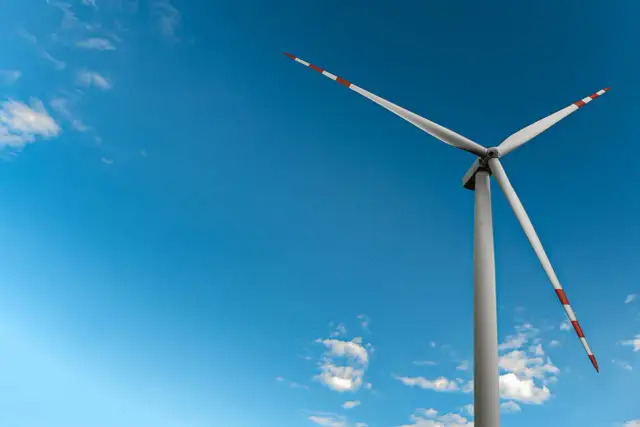 Mała elektrownia wiatrowa z białymi łopatami i czerwono-białymi końcówkami na tle błękitnego nieba z chmurami.