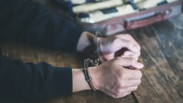 Ręce w kajdankach na drewnianym stole, w tle walizka z paczkami.