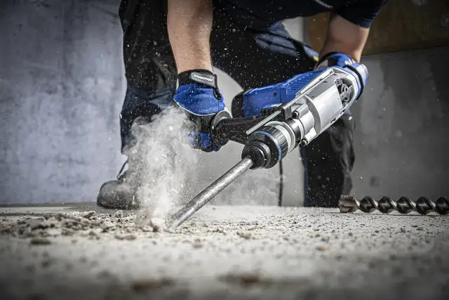 Młotowiertarka z dłutem w betonie, pokazująca jak działa udar w wiertarce. Pył wzbija się w powietrze.