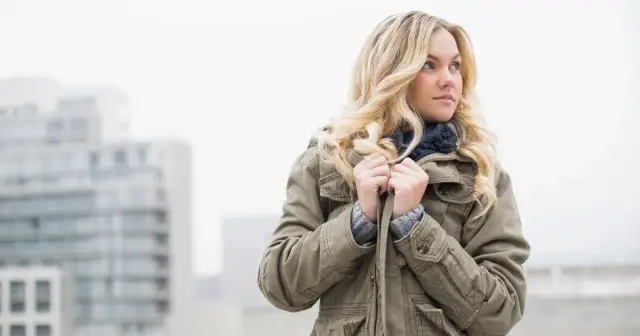 Kobieta w zielonej, ocieplanej parkce z kapturem, otulona szalikiem, pozuje na tle miejskiego krajobrazu.