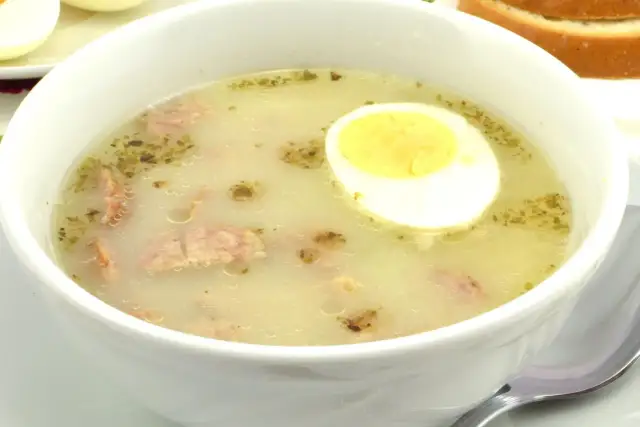Biała miska z zupą, w której widać kawałki kiełbasy i połówkę jajka na twardo.