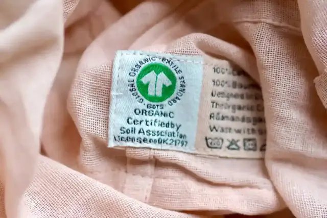 Metka z symbolem GOTS Organic na różowej tkaninie. Informuje o ekologicznym pochodzeniu materiału i zaleca delikatne pranie, bez suszenia w suszarce bębnowej.