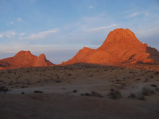 Zachód słońca maluje na pomarańczowo skalne szczyty Spitzkoppe w Namibii. Pustynny krajobraz z suchymi krzewami i ścieżkami.