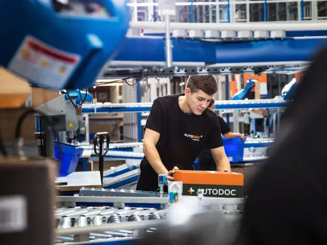 Ein junger Mann in einem AUTODOC-T-Shirt verpackt ein AUTODOC-Paket in einem Lager.