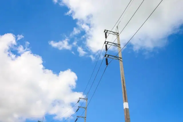 Słupy energetyczne na tle błękitnego nieba z chmurami. Widać przewody i izolatory. Określenie odległości słupa energetycznego od granicy działki jest kluczowe dla bezpieczeństwa.