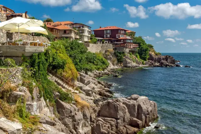 Skaliste wybrzeże z domami na klifie, restauracja z parasolami. Szukasz miejsca, gdzie nad morzem jest najtaniej?