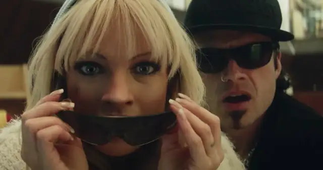 Pam i Tommy w okularach przeciwsłonecznych, ona trzyma je przed ustami, on patrzy z zaskoczeniem.