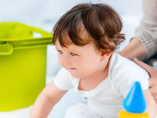 Niemowlę w białej koszulce bawi się zabawkami, obok zielony pojemnik.