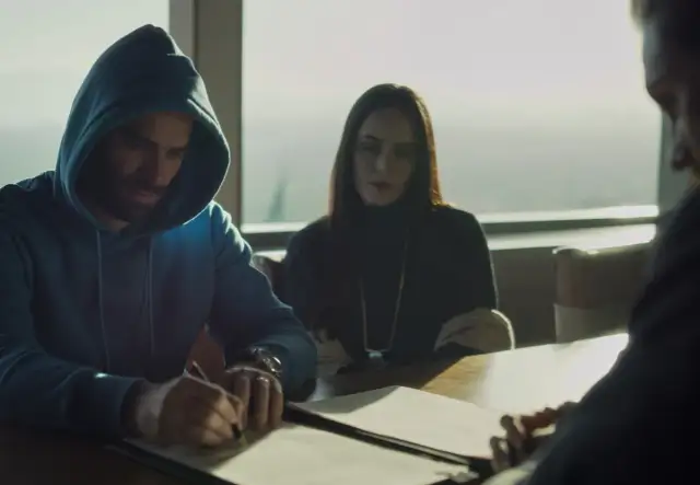 Mężczyzna w niebieskiej bluzie z kapturem podpisuje dokumenty. Obok siedzi kobieta, a z drugiej strony widać fragment twarzy kolejnej osoby. Czy to on wie, kto zabił Sarę?