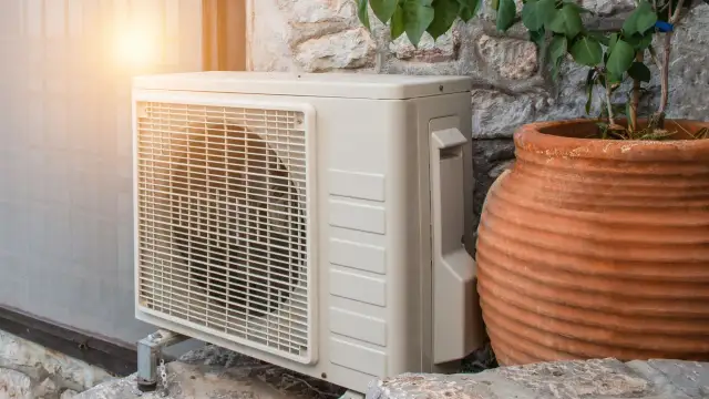 Jednostka zewnętrzna pompy ciepła, obok glinianej donicy z rośliną, w ciepłym świetle słońca. Czas na odpoczynek, kiedy pompa ciepła się wyłącza.