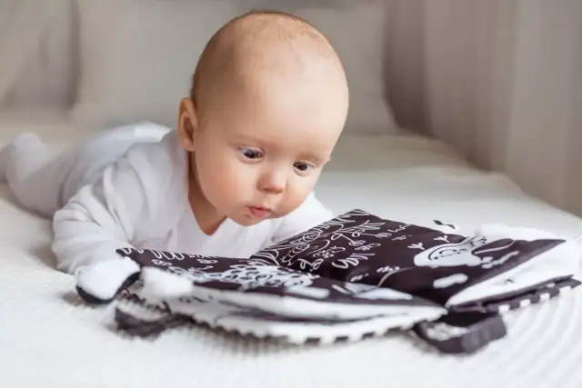 Niemowlę w białym body leży na brzuchu i ogląda czarno-białą książeczkę sensoryczną.
