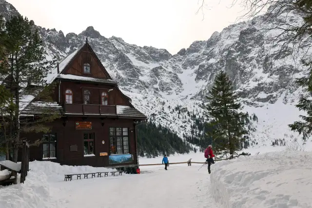 Zimowy krajobraz z drewnianym schroniskiem i ośnieżonymi górami. Turyści wchodzą na teren obiektu. Dowiedz się, ile kosztuje nocleg w schronisku.