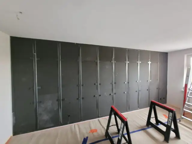 Montaż płyt izolacyjnych na stelażu to pierwszy krok, by wygłuszyć ścianę karton gips. Widać metalowe profile i czarne płyty.