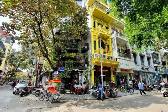 Ulica w Hanoi z żółtą kamienicą, rikszą i skuterami. W tle widać sklepy i restauracje, gdzie można wymienić walutę w Hanoi.