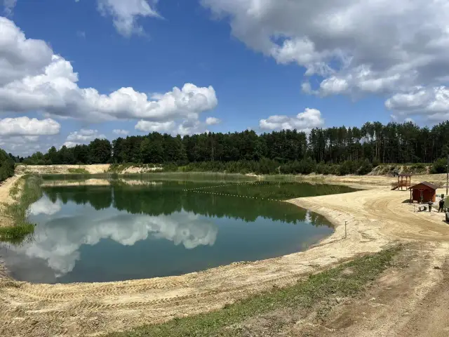 Zbiornik Czarna Sędziszowska z piaszczystą plażą i lasem w tle. Błękitne niebo z białymi chmurami odbija się w spokojnej tafli wody.