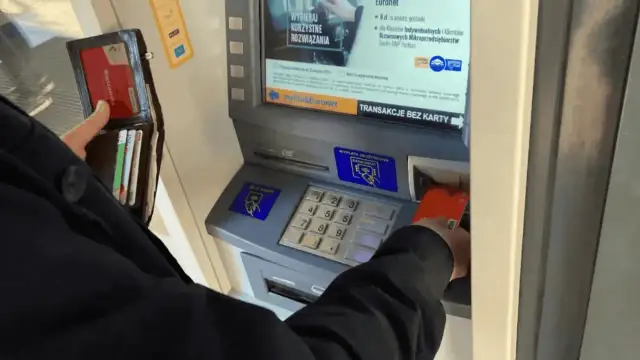 Osoba wkłada kartę do bankomatu, by wypłacić pieniądze. Widoczny portfel z kartami.