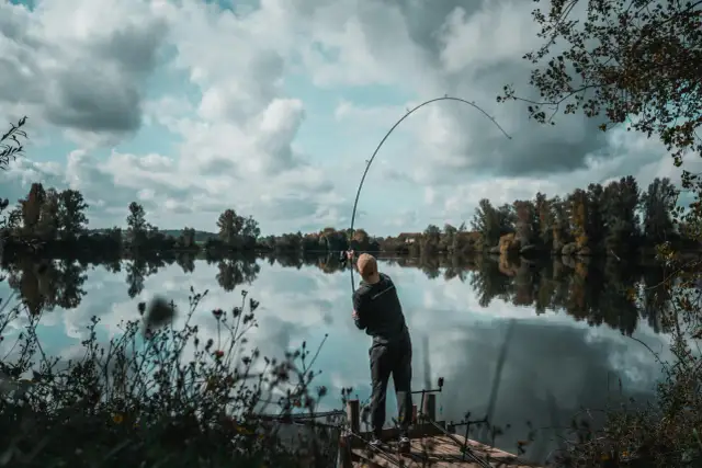 Wędkarz na pomoście łowi ryby na spokojnym jeziorze, z drzewami w tle.