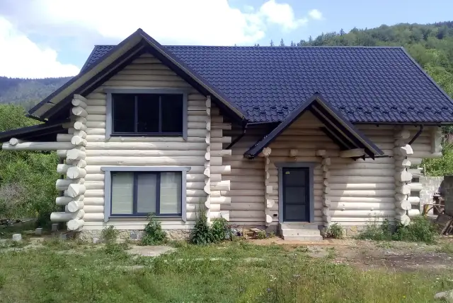 Drewniany dom z bali z ciemnym dachem i oknami, otoczony zielenią.