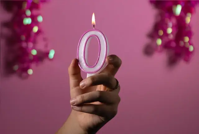 Dłoń trzyma zapaloną świeczkę w kształcie cyfry 0 na różowym tle.