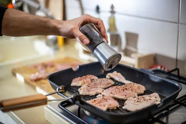 Ręka doprawia pieprzem kawałki kurczaka na grillowej patelni.
