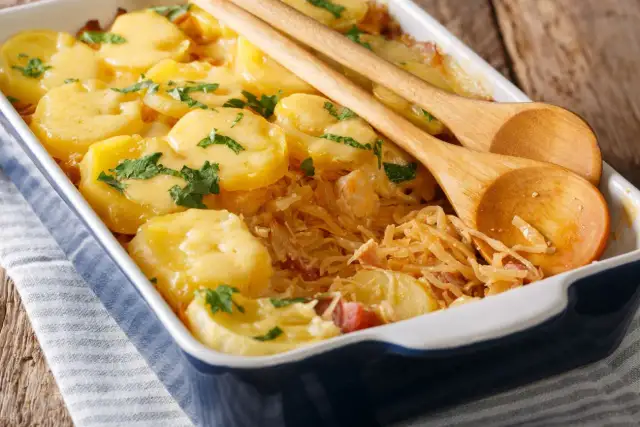 Zapiekanka z ziemniaków i kapusty w naczyniu, ozdobiona natką pietruszki, z drewnianymi łyżkami.