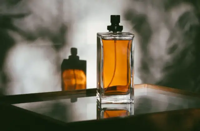 Szklany flakon perfum z bursztynowym płynem i czarną nakrętką, odbijający się w lustrzanej powierzchni.