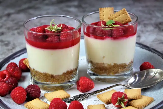 Zwei Gläser mit Himbeer-Dessert, Schicht aus Keksboden, Creme und Himbeersauce, garniert mit frischen Himbeeren und Keksen.
