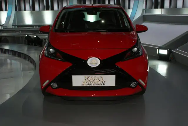 Czerwony samochód z czarnymi akcentami, prezentowany na scenie, z logo "Koło Fortuny" na masce.