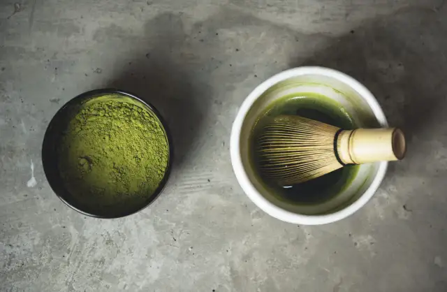 Proszek matcha w czarnej miseczce i zielony napar z bambusową miotełką w białej czarce. Tak przygotujesz herbatę matcha jak pić.