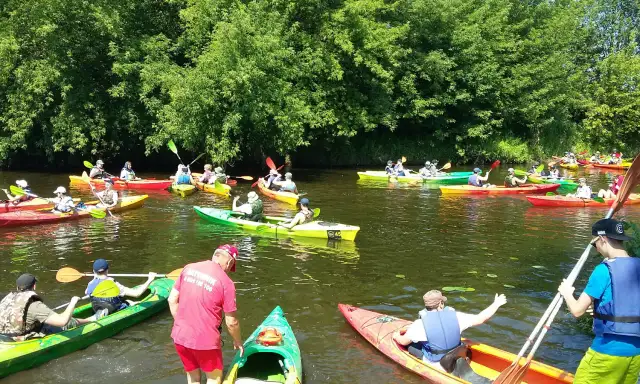 Grupa ludzi na kajakach płynie po rzece, otoczonej zielenią.