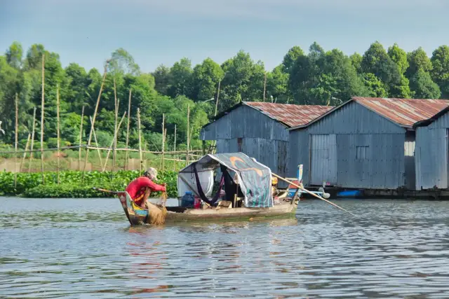 Rybak w łodzi z siecią na tle domów na wodzie i zieleni.