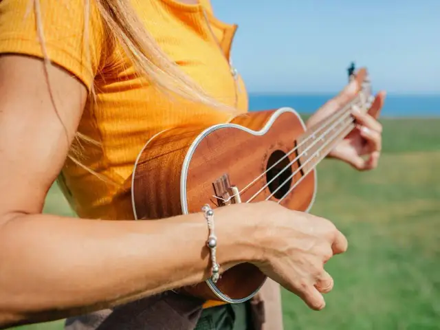 Kobieta w żółtej koszulce gra na ukulele na tle zielonej łąki i morza.