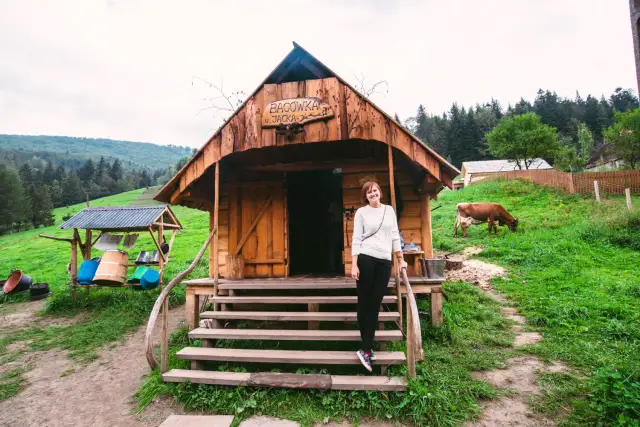 Kobieta przed bacówką u Jancoka, gdzie można kupić oscypki. W tle pasąca się krowa i górski krajobraz.