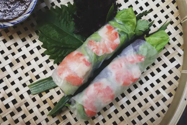 Dwa świeże sajgonki z krewetkami i warzywami, zawinięte w przezroczysty makaron do sajgonek, leżą na bambusowej macie.